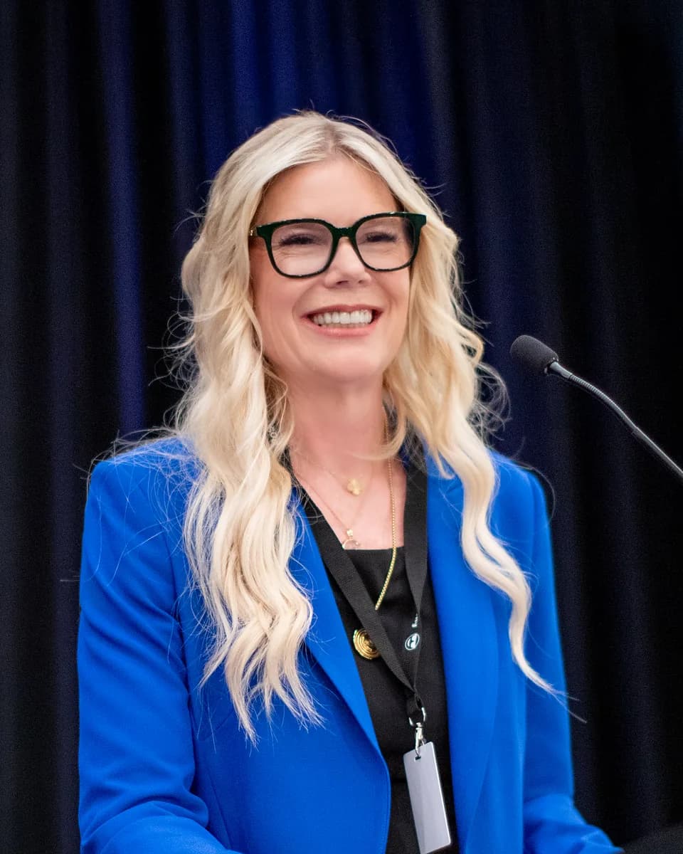 Harmony Vallejo speaking at a lectern in front of a dark blue stage curtain, smiling as she addresses the room, wearing black-framed glasses and a bright blue blazer, with a gooseneck microphone angled toward her