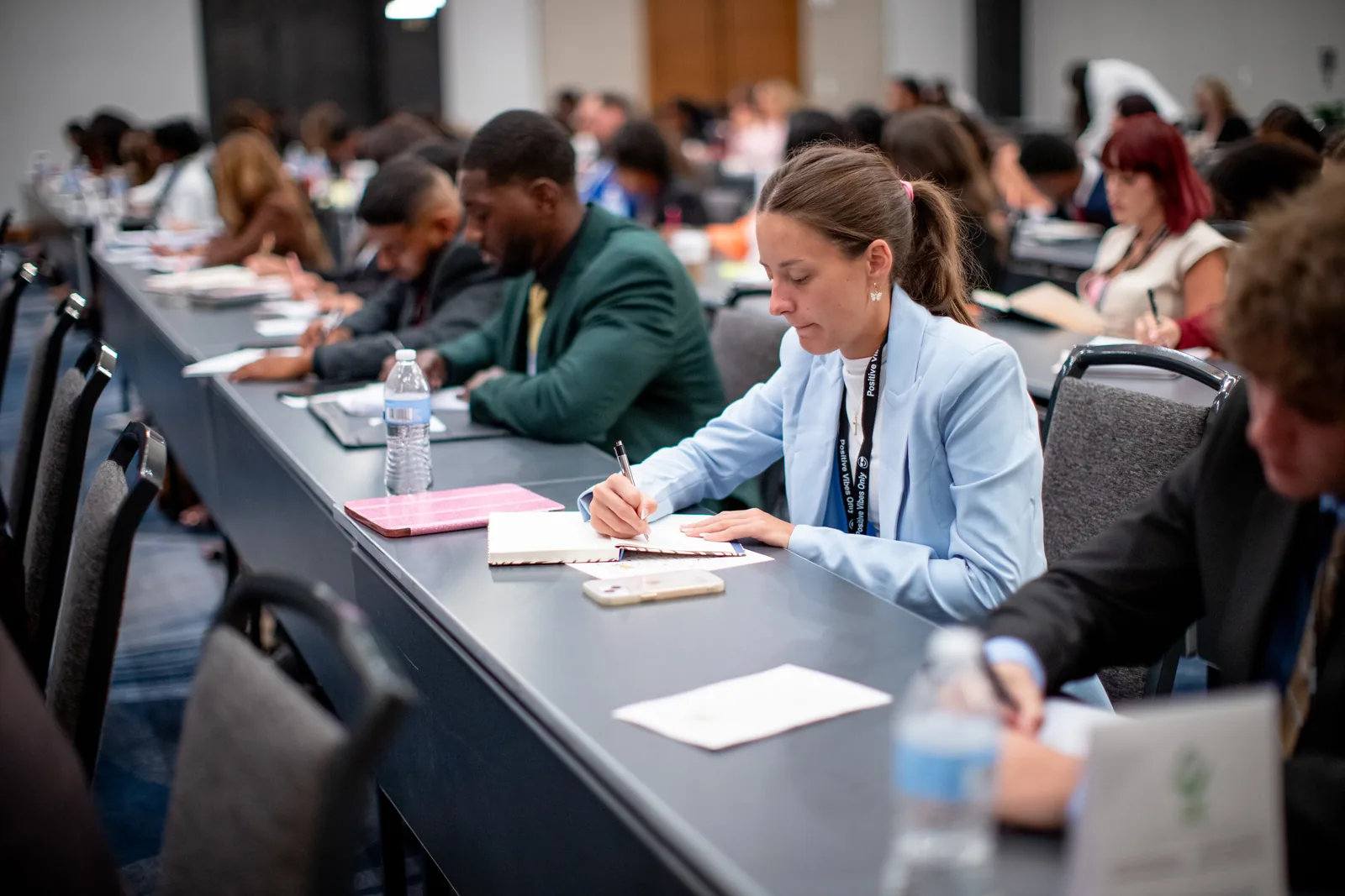 A woman in a light blue blazer writing in a notebook at a long conference table, with rows of other attendees taking notes behind her in a full session room